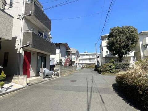 前面道路はゆとりある幅員が確保され、車や自転車での出入りもしやすい道路環境です。周辺には集合住宅や戸建住宅が並び、植栽も見られる落ち着いた街並みが広がっています。現地周辺の道路状況も確認できます。