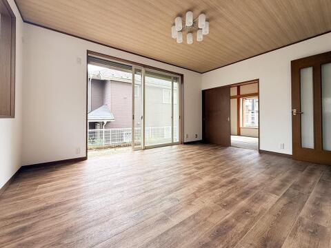 大きな掃き出し窓から自然光が差し込む明るいLDKです。木目調の床と天井が温かみのある空間を演出し、隣接する和室へもスムーズに行き来できます。開口部が広く、庭を身近に感じながら家族で過ごせる住空間です。