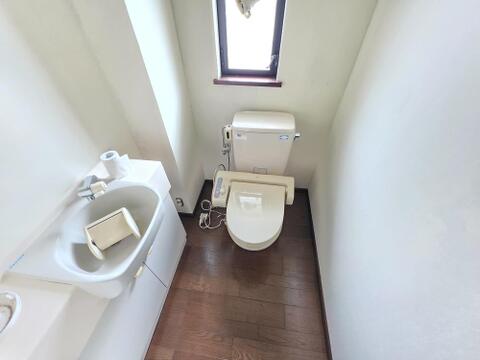 交換前のトイレを撮影しました。便器の横には手洗い器があり、奥の壁には窓が設けられています。図面にはトイレ交換が記載されているため、便器は施工前のものです。手洗い器の位置や室内の形状を確認できます。
