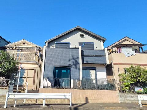 建物外観は、バルコニーの張り出しと外壁の貼り分けが特徴です。道路側には1階・2階ともに大きな窓を設けています。敷地の前面にはフェンスがあり、建物と道路の間に距離を取った配置です。