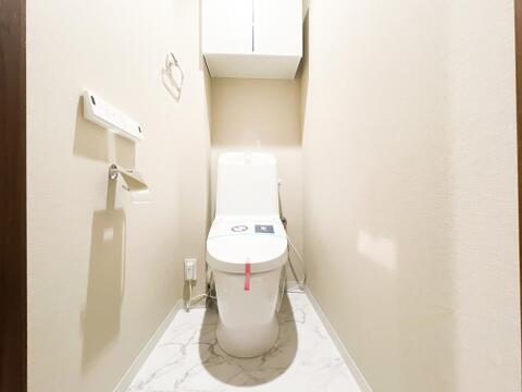 大理石調の床面と温かみのある上品なクロスが調和する、清潔感あふれるトイレ空間。手洗い一体型の便器で省スペースながら機能的であるほか、背部にはトイレットペーパーのストック等に重宝する吊戸棚付きです。