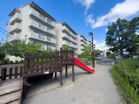 青空の下、広々とした敷地に建つマンション。目の前の公園で、お子様の笑顔を見守り、家族の温かい時間が育まれる住まいです。