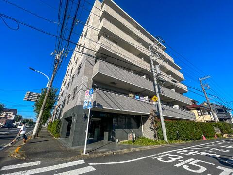 角地に位置するマンションの外観です。グレー系を基調としたタイル張りの外壁で、1階部分にはダークトーンの素材があしらわれており、落ち着いた雰囲気とデザイン性が感じられる佇まいです。
