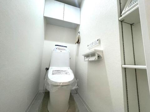 白で統一された清潔感あふれるトイレです。上部の吊り戸棚に加え、右側には可動棚の収納も備えた機能的な空間です。