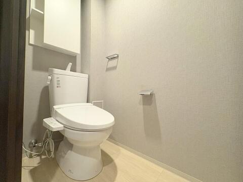 落ち着きのあるグレージュ調の壁紙がおしゃれな雰囲気を与える、清潔感あふれるトイレ空間。上部には便利な吊戸棚が完備されており、トイレットペーパーや備品を隠してすっきり収納できます。
