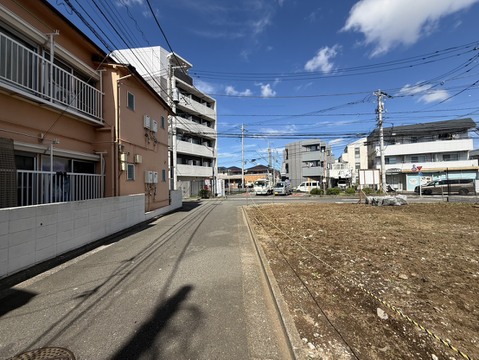 前面道路と隣接する更地、周辺に住宅や建物が見える